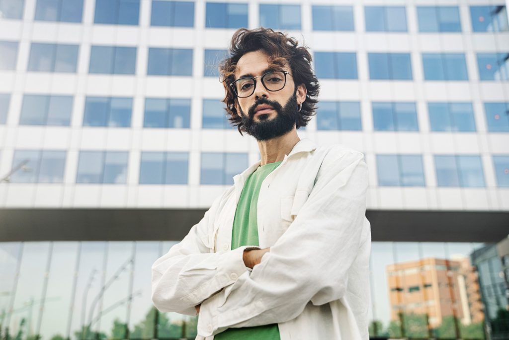 Young confident man with dark hair and beard wearing eyeglasses and standing with arms folded.